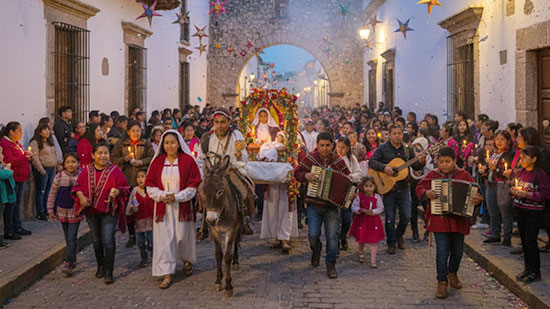 A traditional Mexican Posada procession at dusk, featuring a large crowd carrying candles
