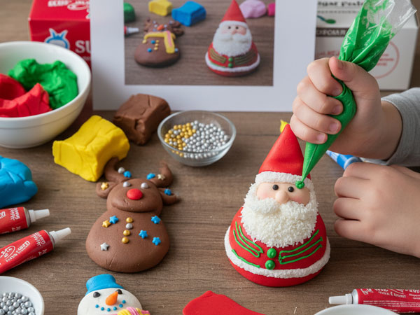 Child hands decorating a fondant Santa Claus figure with green piping frosting, surrounded by other decorated figures (reindeer, snowman)