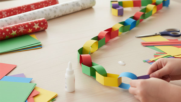 Close-up of hands making a long paper chain garland using strips of colorful construction paper, with glue, scissors, and wrapping paper rolls visible on a wooden table.